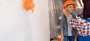 Yeni ev yenileme konsepti. Anne ve oğlu duvarları boyuyor. Anne oğlanın kafasına turuncu kask takar. Birlikte dairelerin tamiri. Pankart.