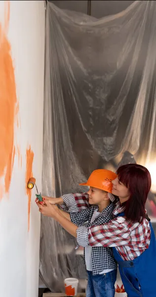 A mãe e o filho estão a pintar uma parede numa casa nova. Uma mulher ensina uma criança a pintar com um rolo enquanto conserta um apartamento. Conceito de renovação de casa. — Fotografia de Stock