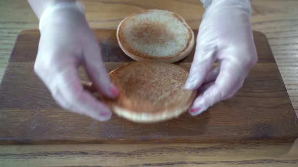 Готуючи бургер в кафе на дерев'яній дошці наливаємо підсмажену булочку з кунжутом з білим соусом. кухарі руки в рукавичках готують гамбургер — стокове відео