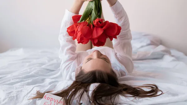 Caucasian girl lying on the bedroom bed with a bouquet of red flowers. tulips in the hotel room in the morning. Dreaming romantic woman birthday — Stock Photo, Image