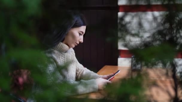 아름다운 갈색의 갈색 머리새는 가을에 비디오 통화를 하는 도시 야외 카페에 앉아 있다. 그 소녀는 휴대폰을 사용하여 의사소통을 합니다. 프로필 뷰 — 비디오