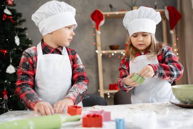 Küçük erkek ve kız kardeş mutfakta Noel kurabiyeleri hazırlıyor, çocuklar hamuru küfle kesiyor, mutfakta pişiriyor. Şenlik arka planı. Boşluğu kopyala
