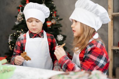 Mutfakta Noel kurabiyesi yiyen küçük mutlu çocuklar, şef üniformalı kardeşler evde yemek pişiriyor, çocuklar ve Noel kurabiyesi. Noel çocuk menüsü.