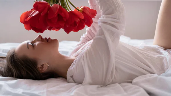 Muchacha joven por la mañana en la cama con un ramo de tulipanes rojos, flores para el día de la mujer, madre día mujer en el dormitorio, primer plano. — Foto de Stock