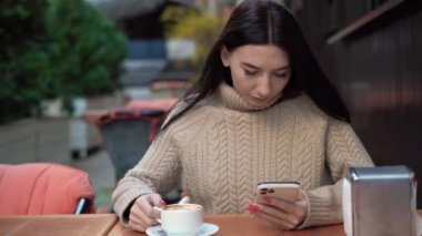 Genç bir kadın bir sokak kafesinde telefonda oturuyor, mesajlara cevap veriyor, sosyal ağlardaki mesajlar, telefon görüşmeleri, çevrimiçi çalışıyor, kapatıyor. Sonbahar
