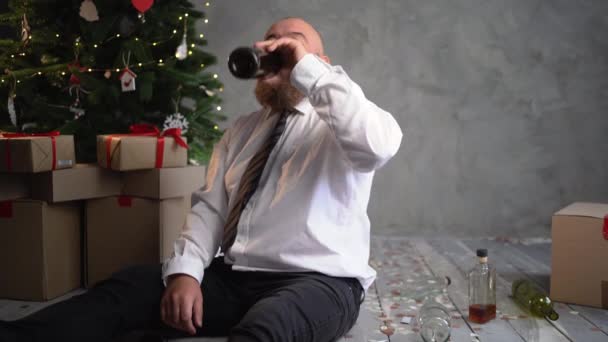 Betrunkene männliche Büroangestellte feiert eine Weihnachtsfeier. bärtiger Geschäftsmann mit Weihnachtsmütze, der Wein aus einer Flasche trinkt, während er auf dem Boden sitzt. — Stockvideo