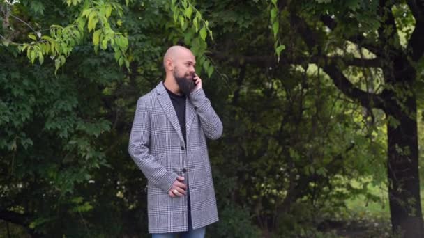 Elegante hombre de negocios barbudo en el parque hablando por un teléfono celular. Un empresario con abrigo recibe una llamada de un colega. Trabajando al aire libre. Trabajo remoto. — Vídeo de stock