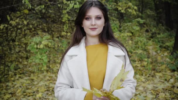 ฤดูใบไม้ร่วงในป่า หญิงสาวยืนอยู่ในสวนยิ้มปกคลุมใบหน้าของเธอด้วยช่อดอกไม้ของใบสีเหลืองที่ลดลงใกล้ชิด เสรีภาพในธรรมชาติ . — วีดีโอสต็อก