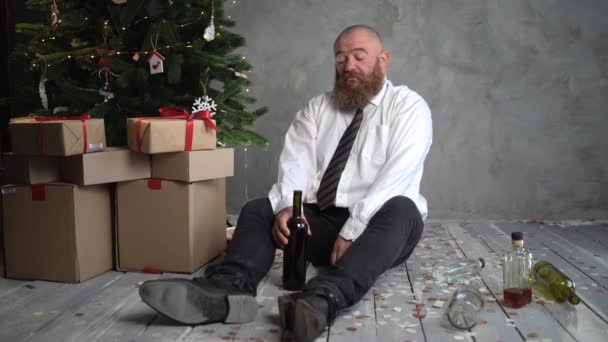 Manhã depois de uma festa corporativa de Natal ou Ano Novo, um homem careca e barbudo bêbado senta-se no chão perto de uma árvore de Natal e bebe vinho de uma garrafa. — Vídeo de Stock