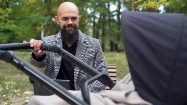 En skallig och skäggig vit man sitter på en bänk i parken och skakar en barnvagn med ett barn. — Stockvideo