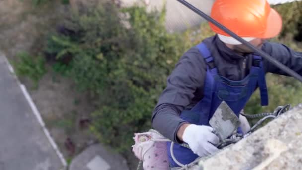 High-altitude work on houses, a Caucasian male worker dressed in a helmet hangs on a rope and puts putty on the house. Renovation work. — Stock Video