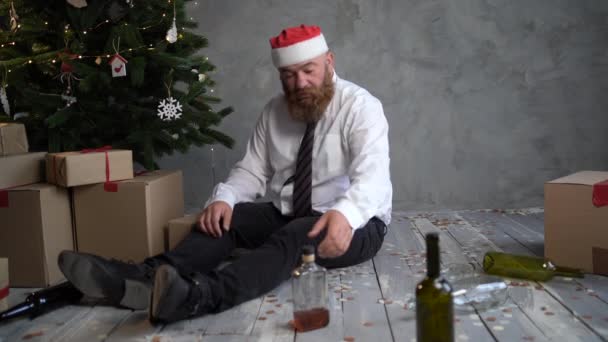 早上，一个喝醉了的大胡子男人在圣诞树旁醒来，他戴着一顶圣诞老人帽，打开一个盒子，盒子里装着礼物，盒子里亮着亮光. — 图库视频影像