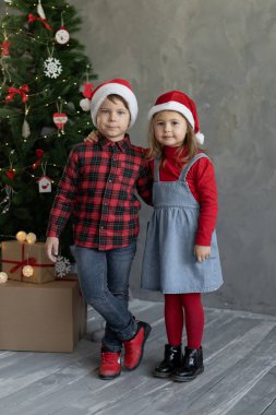 Noel ağacının yanındaki evde yakın çekim çocukları, Noel Baba şapkalı bir kızla odada bir oğlan, mutlu tatiller, çocukluk, yeni yılda aile ilişkileri. Ev Noel konsepti.