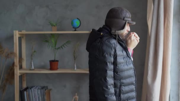 Problemas de aquecimento no inverno. Um jovem caucasiano caminha em casa usando um chapéu e um casaco respirando em suas mãos tentando aquecê-los. — Vídeo de Stock