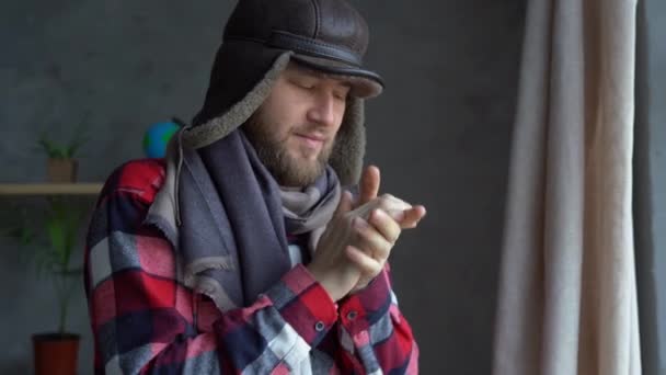 Проблемы с отоплением зимой. Кавказский бородатый мужчина стоит дома у окна в теплой шляпе и шарфе дышит на руках, пытаясь согреться.. — стоковое видео