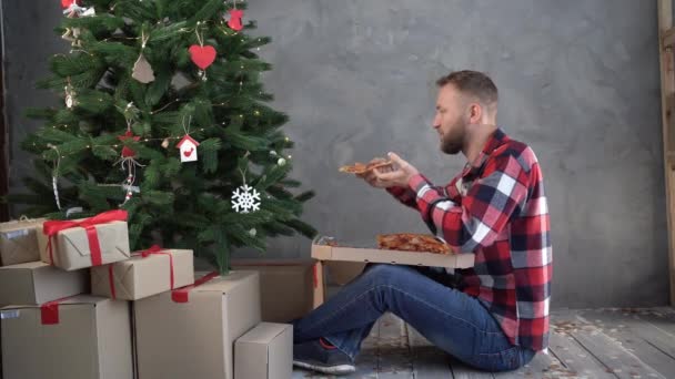 Entrega de pizza para Navidad, hombre barbudo cenando en casa cerca del árbol de Navidad, mordiendo un pedazo de deliciosa pizza italiana, concepto de entrega de comida de vacaciones — Vídeos de Stock