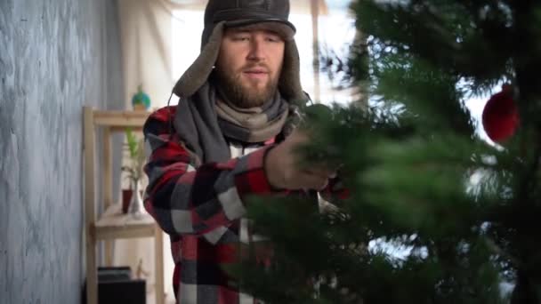 Een man met een hoed versiert een kerstboom thuis, een droevige bebaarde man in een koud huis op kerstavond, verwarmingsproblemen, bevriezing — Stockvideo