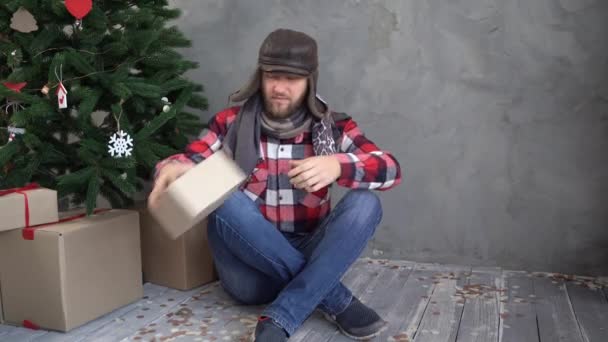 Beau barbu tenant un cadeau du Nouvel An et joyeux Noël émotionnellement, assis près d'un arbre de Noël sur le sol, froid à la maison — Video