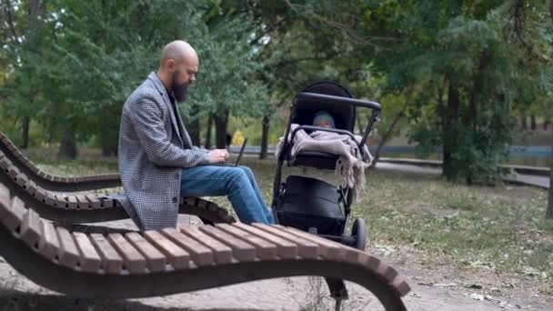 Bello giovane uomo d'affari che lavora a distanza nel parco pubblico con computer portatile che cammina con il bambino in passeggino all'aperto — Video Stock