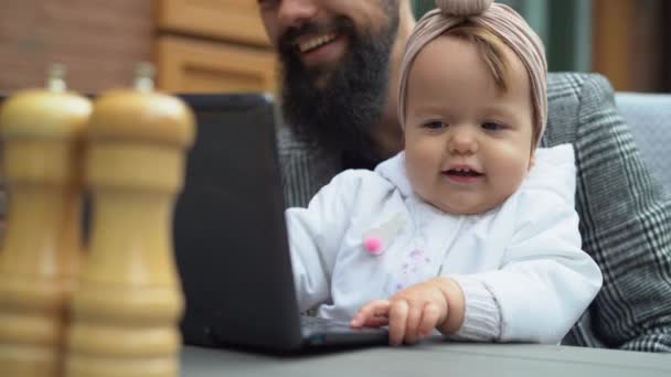 Pai e filha sentados em um café de rua, trabalhando em um laptop on-line, brincando com uma criança em uma mesa de lanchonete, infância feliz — Vídeo de Stock