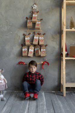 Kot pantolonlu çocuk, duvarda asılı duran takvimin altında yerde oturuyor, çocuklar için şekerli el yapımı kağıt torbalar, evde Noel arifesinde bir çocuk, geleneksel hediye konsepti.
