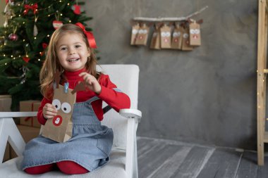 Noel hediyesi konsepti. Elinde takvim paketi olan mutlu küçük bir kız. Evinde bir koltukta oturuyor. Odadaki duvarın yanında bir Noel ağacı. Neşeli Noel arkaplanı