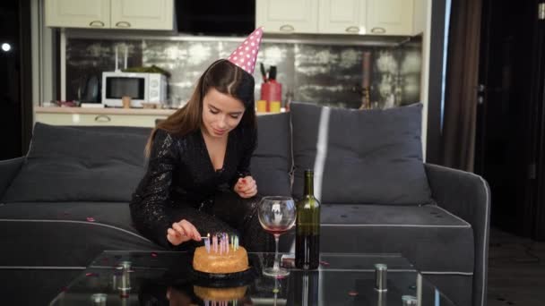 Een jonge blanke dame zit thuis op de bank op haar hoofd met een verjaardagskapje en steekt feestelijke kaarsen in de taart. Huis voor het vakantie concept. — Stockvideo