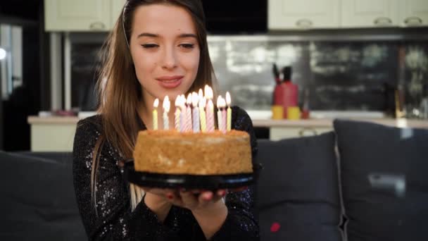 Молода кавказька жінка сидить вдома на дивані на голові, вдягнена в шапку з днем народження і тримає торт зі свічками. засяє бажання відкрити очі і погасити свічки.. — стокове відео