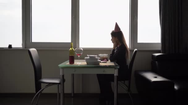 Mladá žena sedí sama u stolu o svých narozeninách. slavnostní klobouk na hlavě. Dáma se cítí znuděná a osamělá. Láhev vína a narozeninový dort. Smutný a osamělý narozeninový koncept. — Stock video