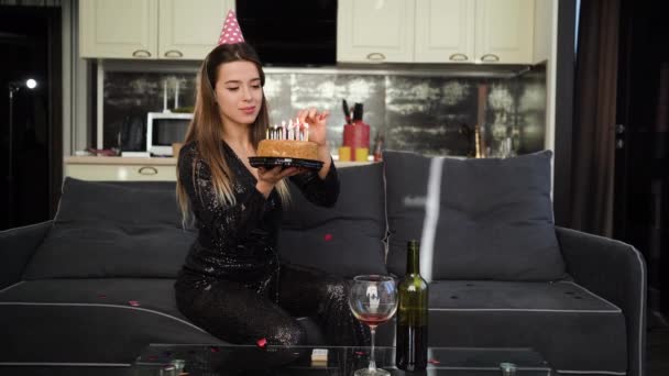 Una joven mujer caucásica se sienta en casa en un sofá en su cabeza con una gorra de cumpleaños y sostiene un pastel de cumpleaños en sus manos y enciende velas. concepto de preparación para unas vacaciones felices. — Vídeos de Stock