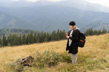 Doğada tatilde olan bir kız gezgin ve akıllı bir telefondan online bir rota haritası çizen bir turist kız, bir dağ manzarasının arka planında tatilin tadını çıkaran bir cep telefonu kullanıyor.