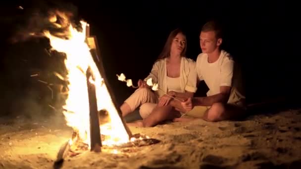 Una giovane coppia uomo e donna si riposano accanto al fuoco, seduti sulla spiaggia sabbiosa che si abbracciano e si divertono a parlare e friggere marshmallow. — Video Stock