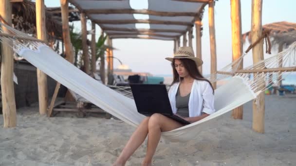 Счастливая молодая внештатная предпринимательница в шляпе сидит на гамаке с современным ноутбуком и работает удаленно на берегу тропического острова во время летних каникул. копировальное пространство. — стоковое видео