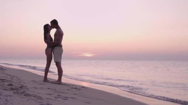 Frühmorgens im Morgengrauen oder abends bei Sonnenuntergang steht ein junges verliebtes Paar, ein Mann und eine Frau, am Meer und küsst sich leidenschaftlich. Mann und Frau während eines Familienurlaubs im Resort. — Stockvideo