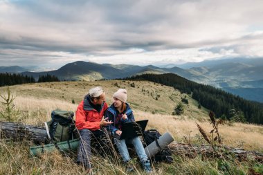 Adam ve kız yürüyüşçüler bir dağ ormanında oturup dizüstü bilgisayara bakıyorlar. Bağımsız çalışanlar dağlarda yürüyüş yaparken çalışırlar. Baner. Boşluğu kopyala.