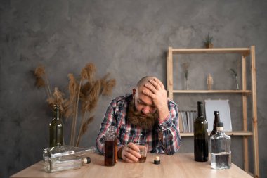 Yetişkin sakallı bir adam masada oturmuş evde bir şişe alkol içiyor üzgün, alkolizm, eliyle başını tutuyor. Rehabilitasyon suistimalinin ve iyileşme sorununun belirtileri. Boşluğu kopyala.