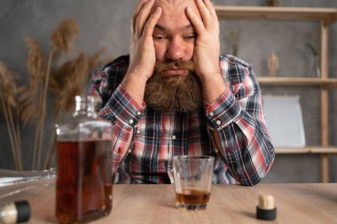 Alkolizm konsepti. Genç adam çok fazla alkol içiyor. Evde gündelik kıyafetlerle oturuyor, başını konyak ve viski şişelerinin üzerine eğiyor. Bağımlılık ve hastalık. Boşluğu kopyala.