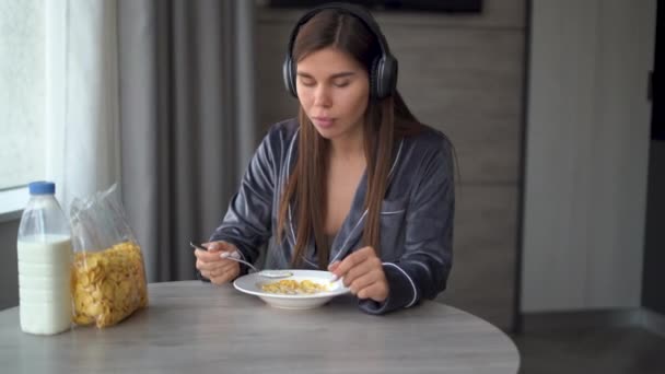 Młoda biała kobieta siedzi rano w piżamie, sama przy stole, jedząc płatki kukurydziane z mlekiem na śniadanie. Słuchawki są noszone, słuchając muzyki i tańcząc w domu. — Wideo stockowe
