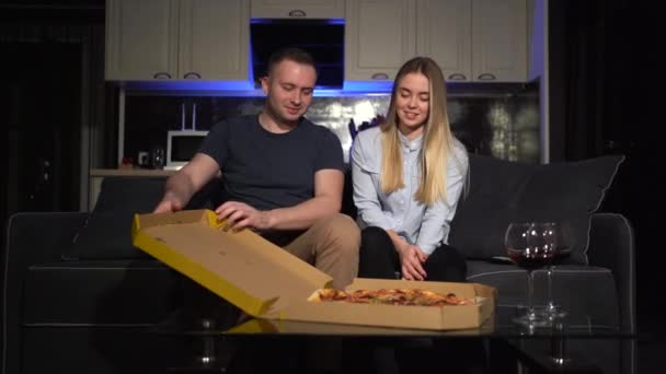 Stând pe canapea acasă seara. Cuplul tânăr petrece timp împreună. Bărbatul deschide cutia de pizza și ia o mușcătură pentru el însuși.. — Videoclip de stoc