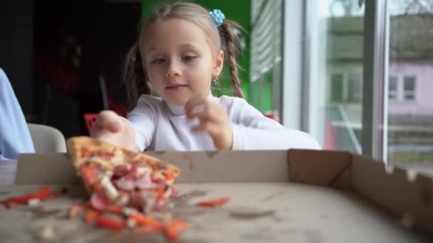 Enfant affamé fille 5-6 ans manger de la pizza dans un restaurant tend la main et sort le dernier morceau de pizza de la boîte. La famille s'amuse ensemble. — Video