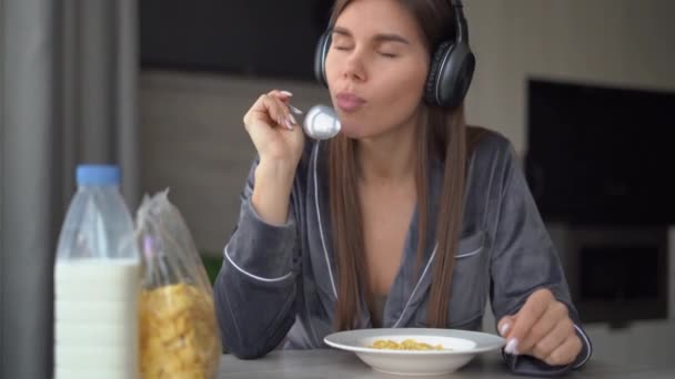 Portret białej kobiety siedzącej rano przy stole w piżamie, jedzącej płatki kukurydziane z mlekiem, noszącej słuchawki na głowie, słuchającej muzyki i tańczącej. — Wideo stockowe