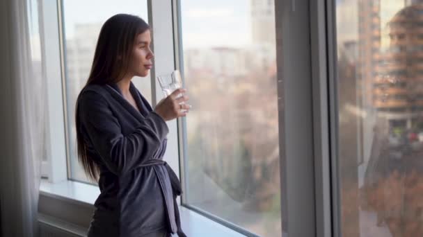 Jeune belle femme caucasienne vêtue de pyjama tôt le matin se tient près de la fenêtre tenant un verre d'eau propre dans ses mains et reconstitue les réserves de h2o dans le corps. — Video