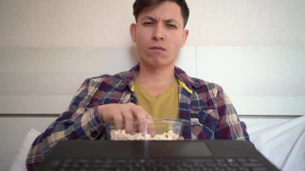 Jeune homme caucasien assis à la maison sur le canapé en vêtements décontractés pendant la journée manger du pop-corn et regarder un film. Émotion de surprise et examine attentivement quelque chose et s'approche de la webcam. — Video
