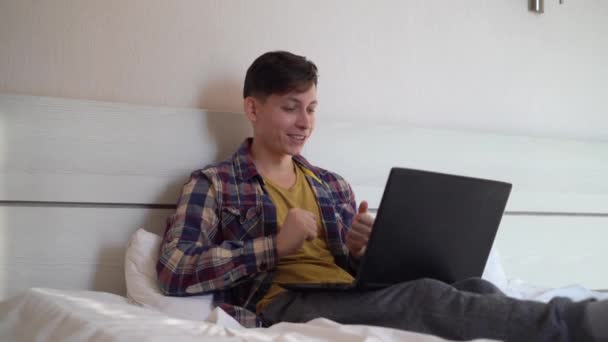 Молодой кавказский мужчина, сидящий дома на кровати в повседневной одежде и общающийся по видеосвязи с помощью ноутбука. C — стоковое видео