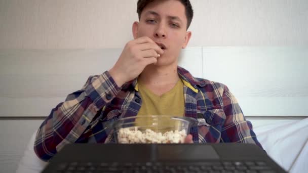 Joven hombre caucásico sentado en casa en el sofá con ropa casual durante el día comiendo palomitas de maíz y viendo una película. calmar la expresión no emocional en su cara. — Vídeo de stock