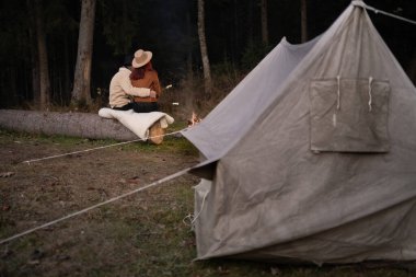 Sonbahar ya da ilkbaharda güzel bir gecenin keyfini çıkarırken, kamp yerindeki parlak, yanan bir kamp ateşinin yanında şapkalı turistlerin arka plandaki görüntüsü. Boşluğu kopyala.