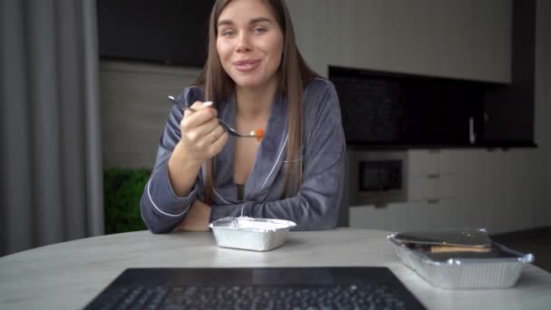 Großaufnahme Porträt vom Bildschirm. Glückliche junge Frau sitzt zu Hause am Tisch in der Küche, frühstückt mit Salat mit Einweggeschirr und macht einen Videoanruf von ihrem Laptop. Essensausgabe — Stockvideo