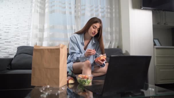 Ein Mädchen während des Abendessens telefoniert mit einem Laptop und sitzt in Hauskleidung auf der Couch. Ohne Gesicht. Hauszustellung. Konzept für gesunde Ernährung. — Stockvideo