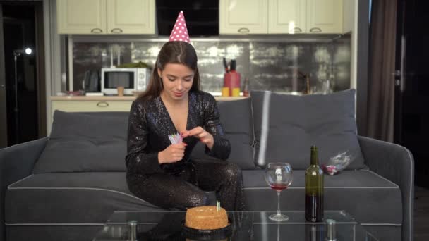 Una joven hermosa dama caucásica está sentada en casa en el sofá en su cumpleaños inserta velas en el pastel de cumpleaños. Una mujer en un sombrero de fiesta. — Vídeos de Stock