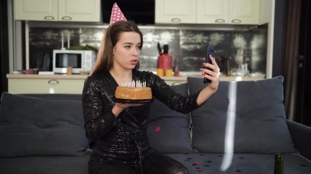 One young Caucasian woman is sitting at home on the couch on her birthday wearing a festive cap and is broadcasting live using her smartphone. Online sad party concept. — Stock Video